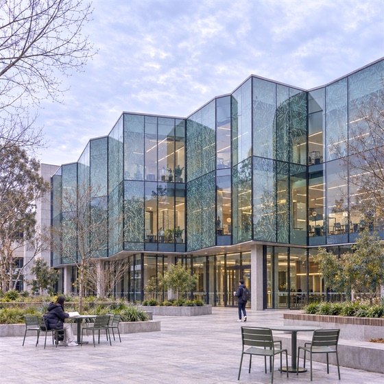 维多利亚大学 Footscray Park 校区：维多利亚大学 Footscray Park P 楼南庭院的冬季阴天示意图，灰绿折面玻璃带抽象蚀刻纹，一名学生阅读，另一名学生步行。